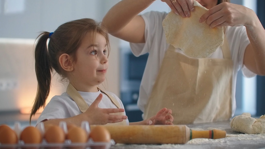 A young girl helps roll out dough in the kitchen while wearing an apron, surrounded by baking ingredients