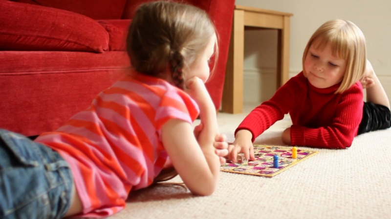 Two young children lie on the floor, focused and smiling as they play a board game together in a cozy living room