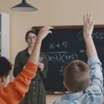 A group of children eagerly raising their hands in front of a blackboard during a classroom activity