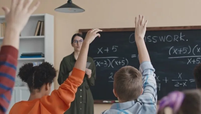 A group of children eagerly raising their hands in front of a blackboard during a classroom activity