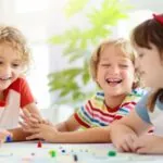 Three young kids laugh and play together around a table, fully engaged in one of several board games that make learning math fun
