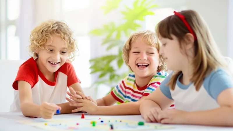 Three young kids laugh and play together around a table, fully engaged in one of several board games that make learning math fun