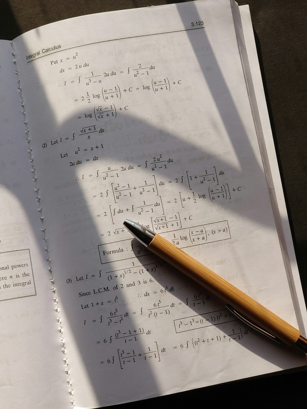 A mathematics textbook open to a page on integral calculus, featuring several worked-out examples involving substitution and logarithmic integration techniques, with a pen resting on the page