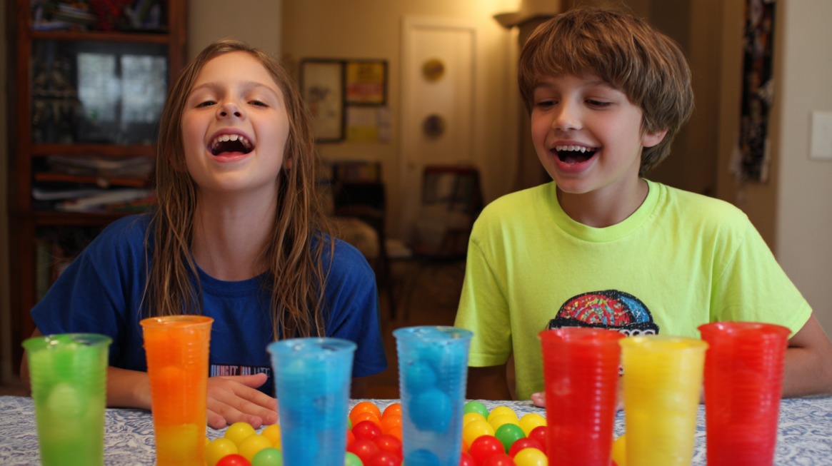 Two smiling kids playing a Minute to Win It game with colorful plastic cups and plastic balls on a table