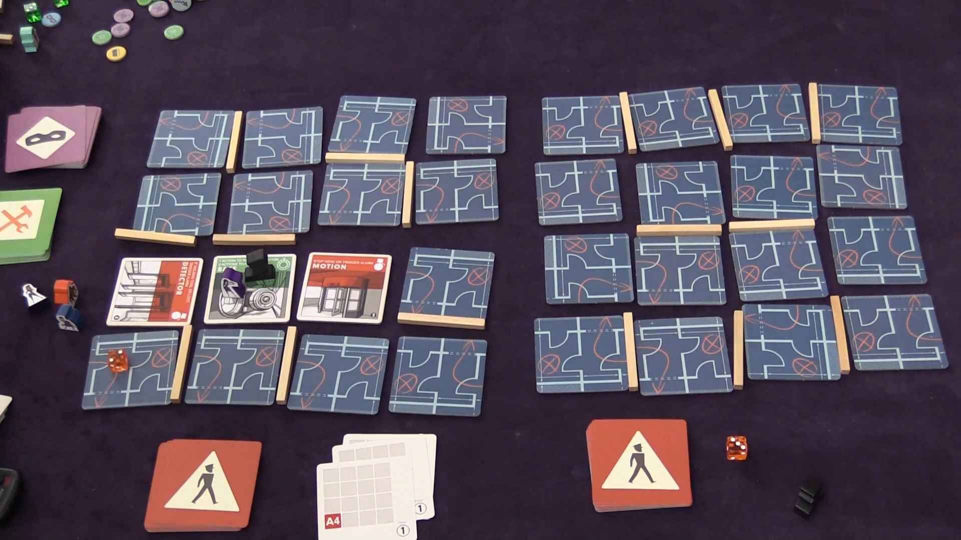 Top-down view of the Burgle Bros board game setup showing face-down tiles arranged in a grid with various cards, tokens, and dice