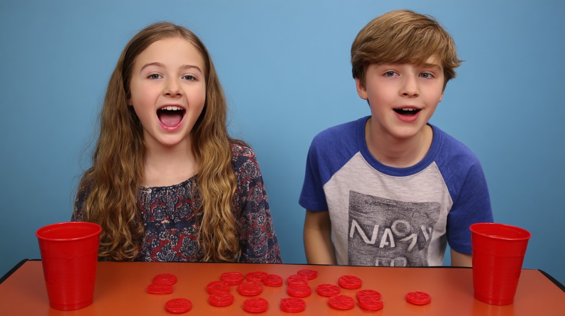 Two smiling kids sitting at a table with red plastic cups and stacked red tokens, ready for a Minute to Win It game