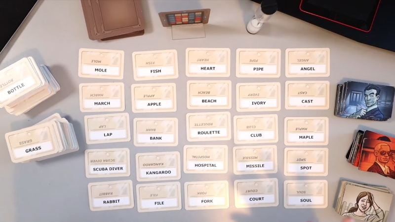A Codenames board game setup with word cards laid out in a grid and stacks of clue cards ready for play