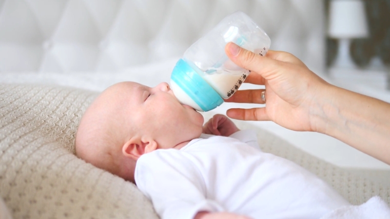 A caregiver feeds a newborn baby with a bottle of infant formula while the baby lies on a soft blanket