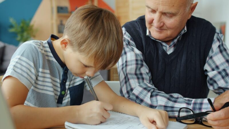 A young boy concentrates on his schoolwork, guided by a supportive grandfather