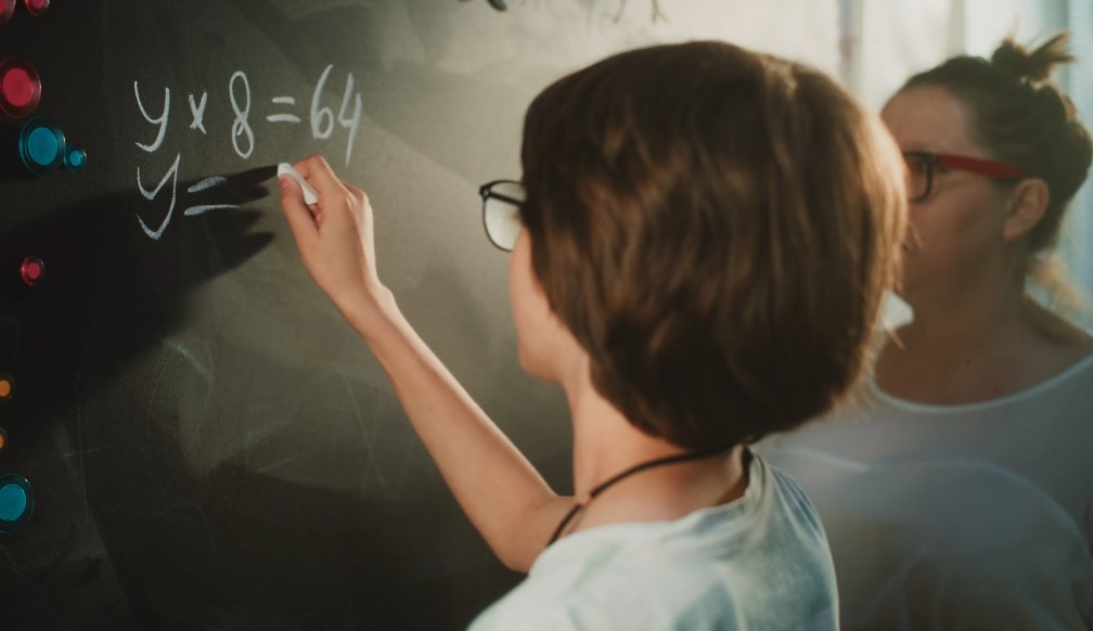 A student solves a math problem on a chalkboard, guided by a teacher