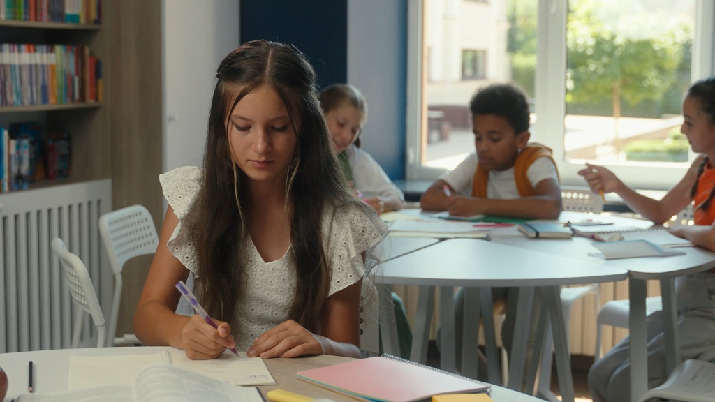 Girl in a white blouse writing notes during study session, surrounded by classmates and colorful notebooks