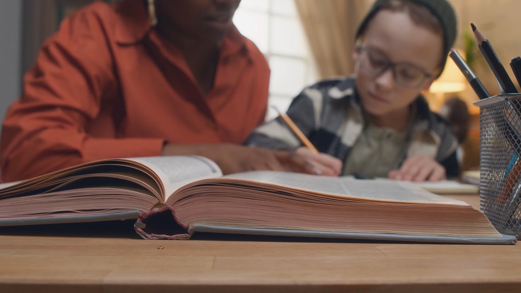 Teacher guiding a young student through a large open textbook at the desk