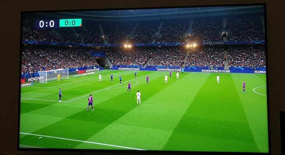 Football match in progress, score 0-0, with players on a green field and a large crowd in the stadium