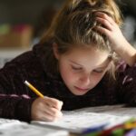 A young girl sits at a table concentrating on homework, writing in a notebook while holding her head with one hand