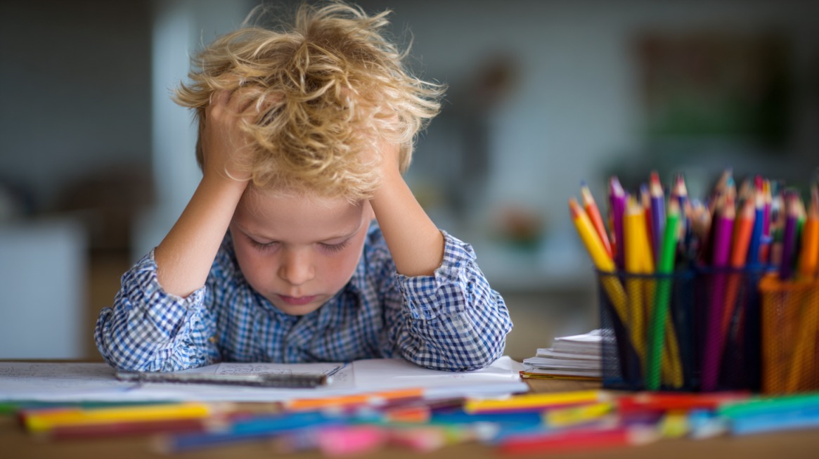 A young child sits at a table holding their head in frustration while trying to complete homework